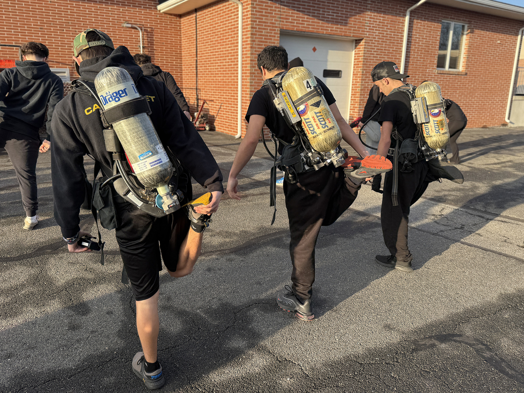 Students in firefighting training gear walk while stretching their legs, each holding one foot behind them, during an outdoor warm-up near a brick building.