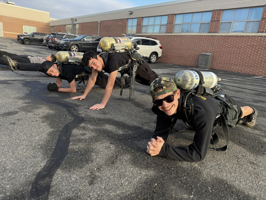 Four students in firefighting gear and air tanks hold plank positions on an outdoor asphalt area, smiling toward the camera, with a brick school building and parked cars in the background.