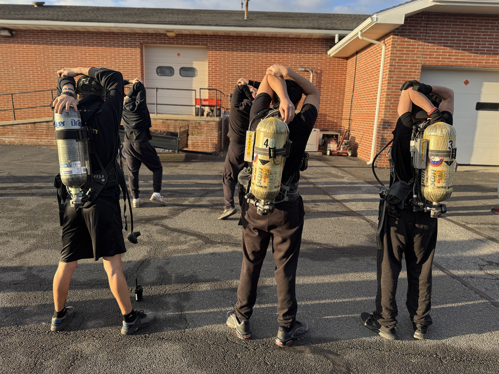 Students wearing firefighting training gear and air tanks perform overhead triceps stretches on an outdoor asphalt surface, with a brick school building in the background.