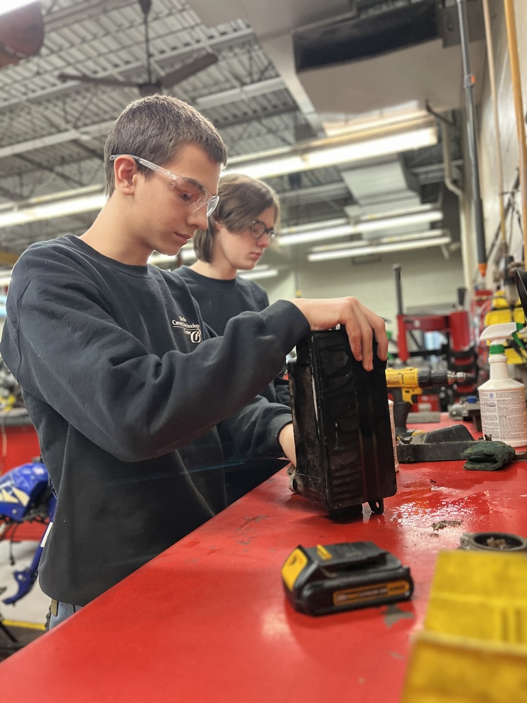 Two students examine a removed mechanical component at a red workbench as part of a hands-on equipment service activity.