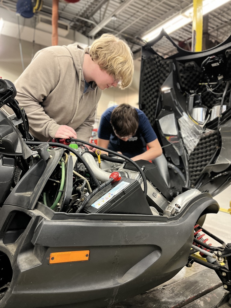 Two students inspect internal components of a snowmobile with body panels removed while working together in a technical training lab.