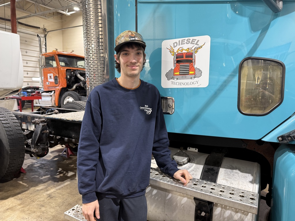 A Diesel Technology student wearing a camo hat and navy BCTC sweatshirt stands in a diesel lab beside a teal semi-truck, with another truck visible in the background.