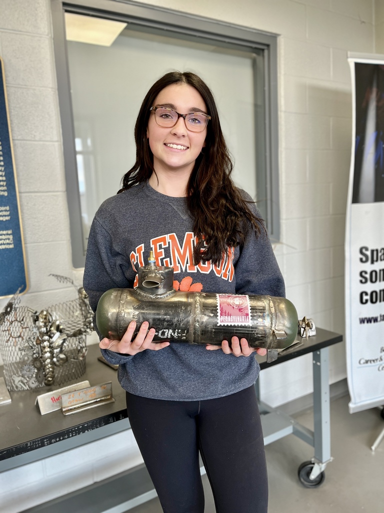 A Berks Career & Technology Center student smiles while holding a metal submersible welding fabrication project in front of a display table inside the school.