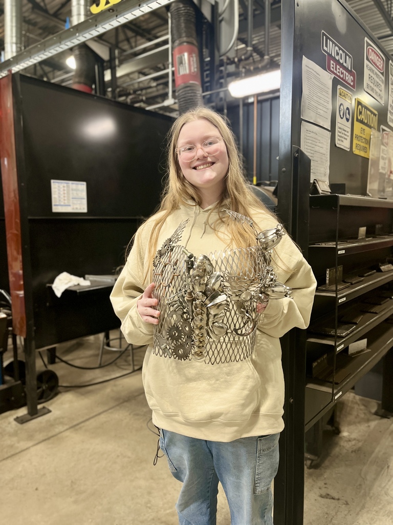 A Berks Career & Technology Center student stands in a welding lab holding a welded metal sculpture made from mesh and metal pieces, with welding booths and equipment behind her.