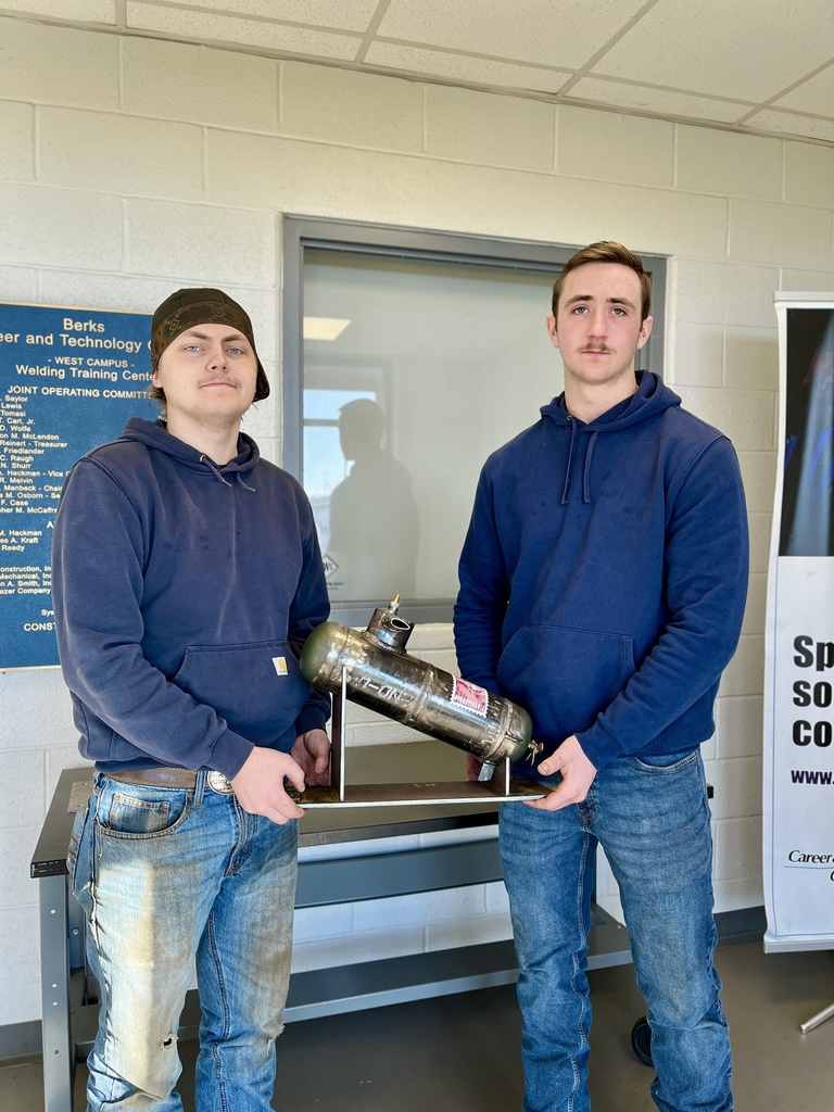 Two Berks Career & Technology Center Welding Technology students stand indoors holding a metal submersible welding fabrication project mounted on a rectangular base.