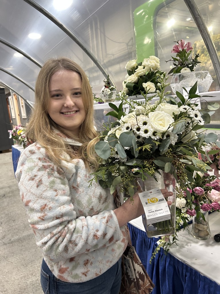 Smiling student holds a large white floral bouquet with greenery at the PA Farm Show; the entry label is visible while floral arrangements are displayed behind her.