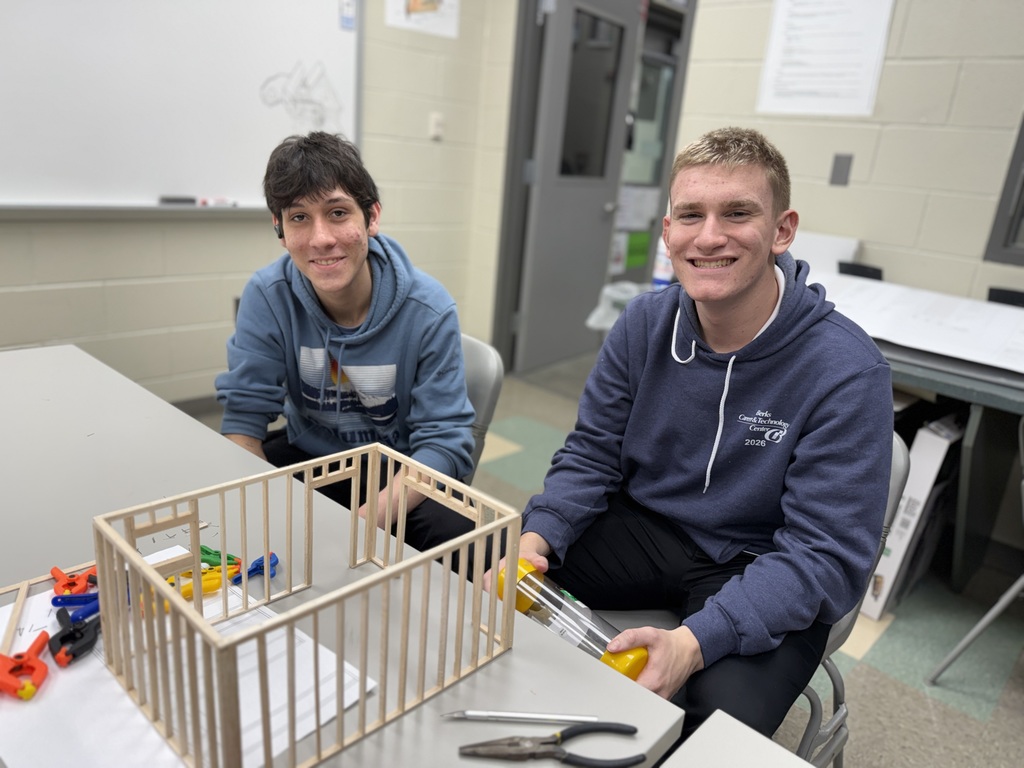 Two Drafting students sit behind a partially built wood-framed shed model on a table with tools and building plans in a classroom.