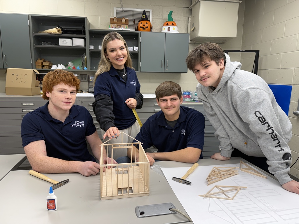 Four Berks Career & Technology Center Drafting students pose in a classroom beside a wood-framed shed model, with roof trusses and printed construction drawings on the table.