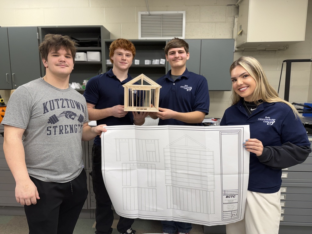 Four Berks Career & Technology Center Drafting students pose in a classroom holding a large shed blueprint while two students display a completed wood-framed shed model.