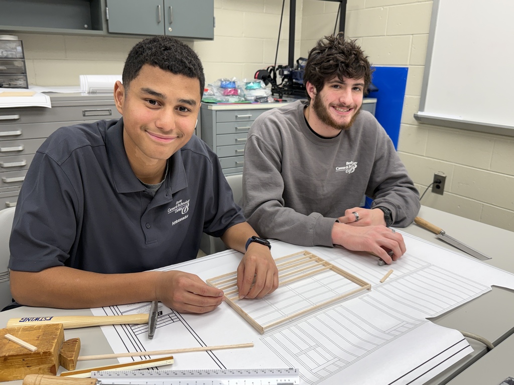 Two Berks Career & Technology Center Drafting students smile while assembling a small wood-framed model on top of large blueprint drawings in a classroom.