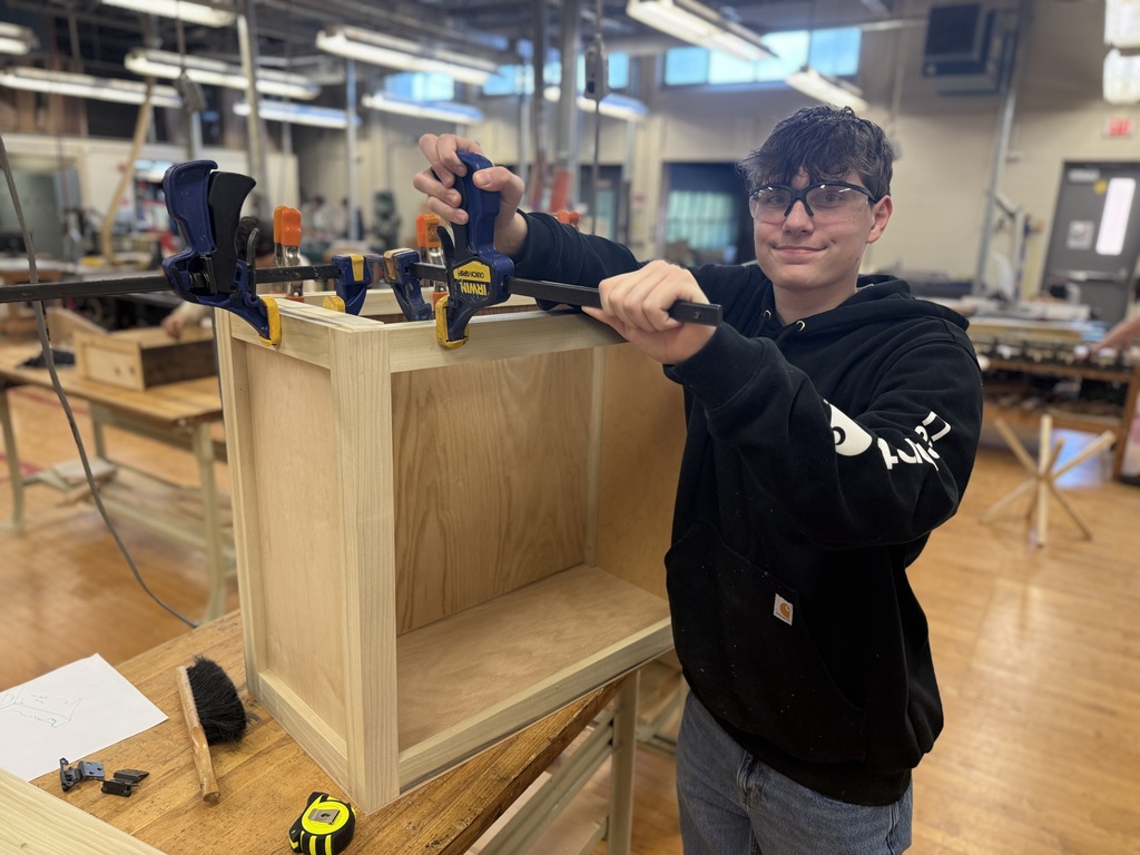 Student in safety glasses uses clamps and a hand tool to assemble a wooden cabinet frame on a workbench in the wood shop.