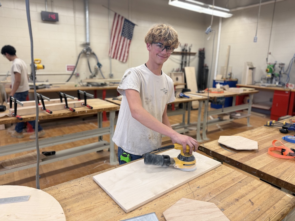 Student wearing safety glasses uses a power sander on a rectangular wood panel at a workbench in the Cabinetry & Wood Technology shop.