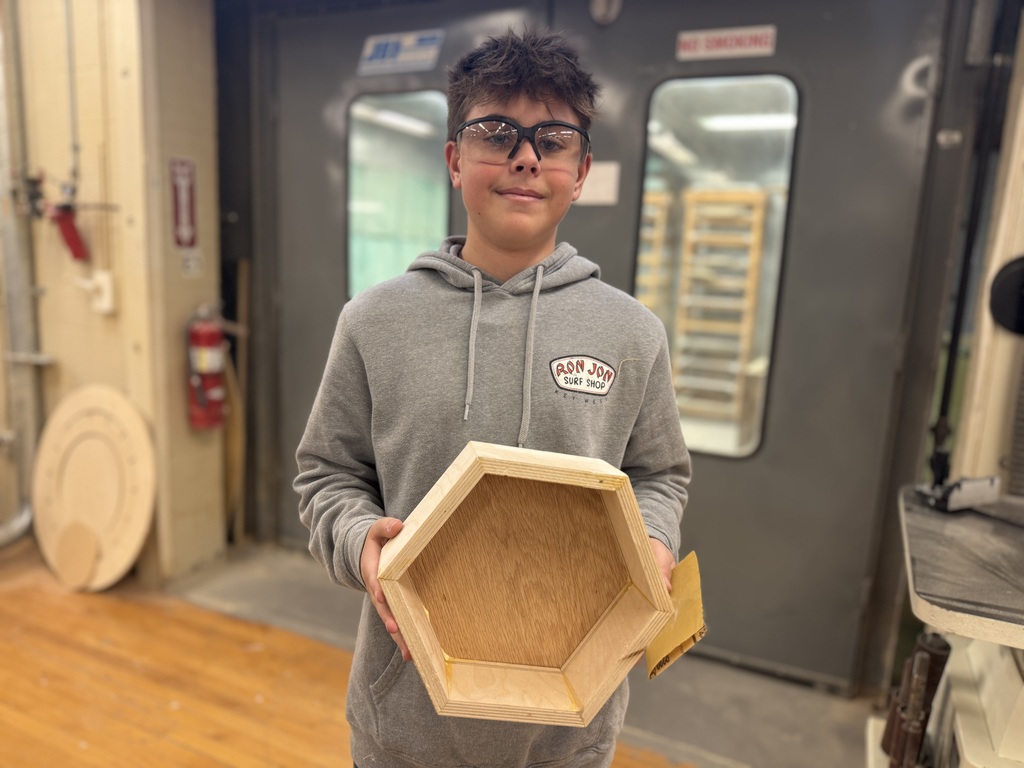 Student wearing safety glasses holds an unfinished octagon-shaped wooden box in front of large shop doors in the woodworking classroom.