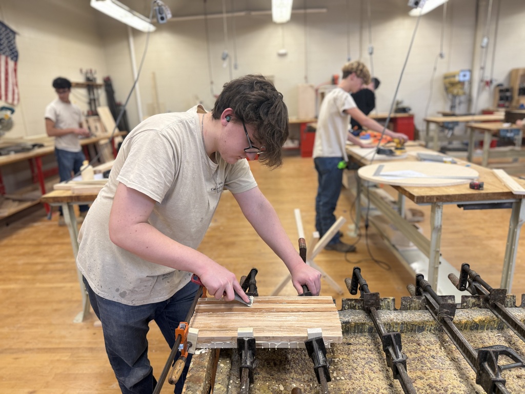 Student sands a glued wood panel clamped to a workbench in a woodworking classroom while other students work in the background.