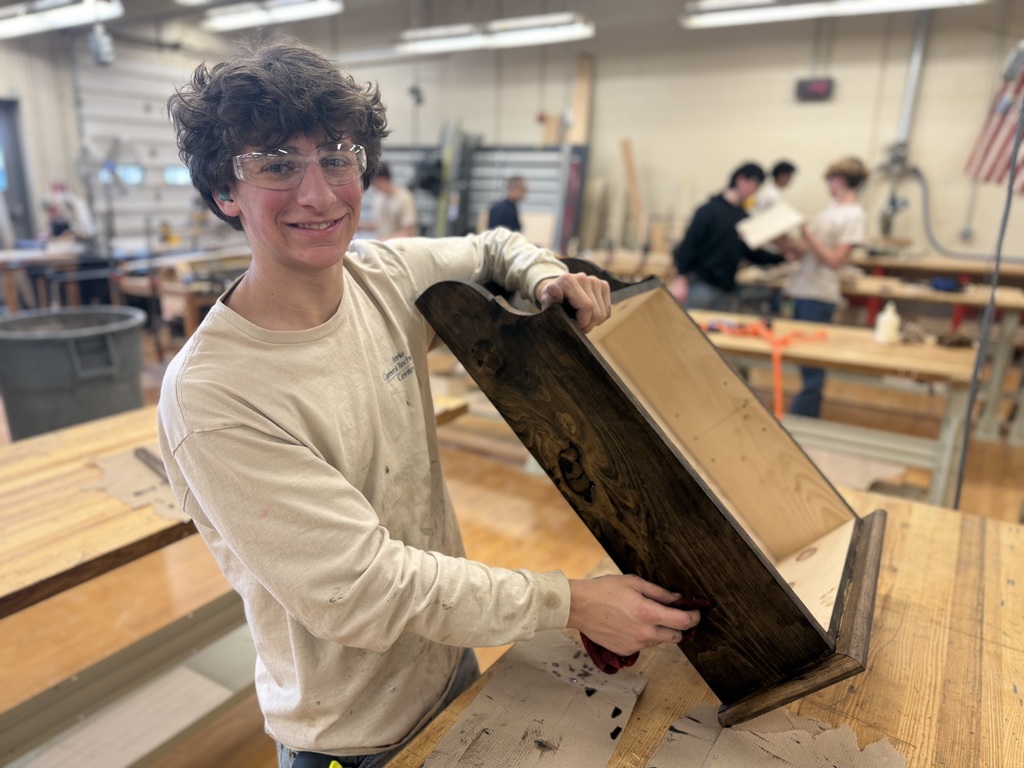 Student wearing safety glasses holds a large wooden project with a dark stained finish inside a woodworking classroom.
