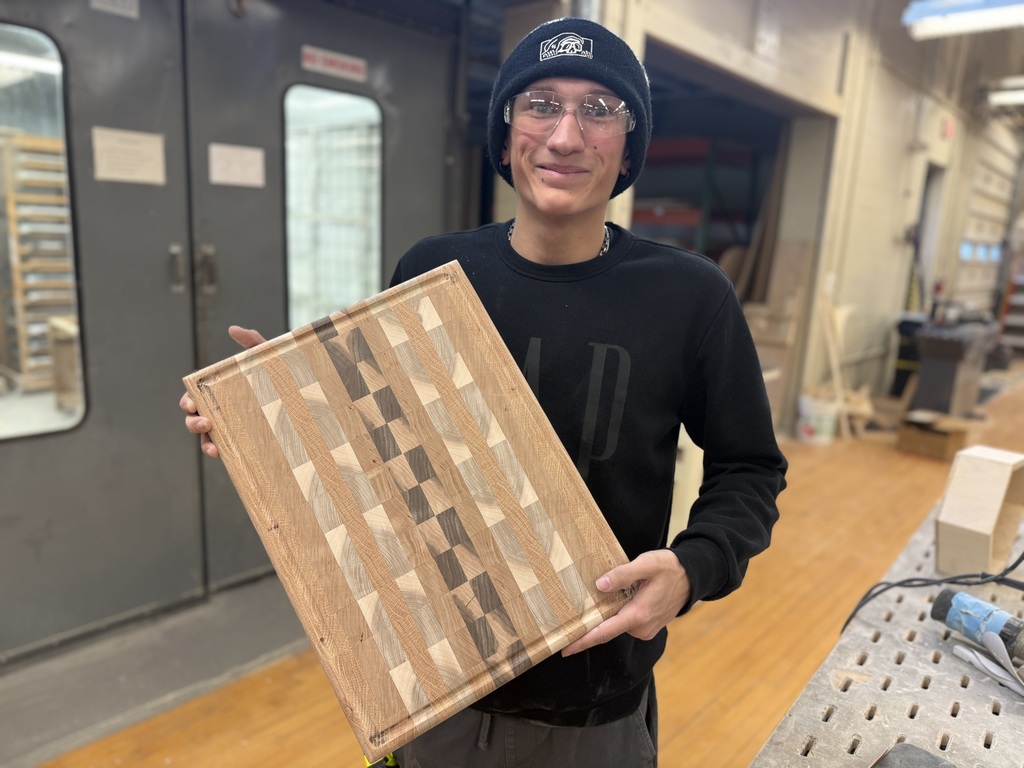 Student wearing safety glasses and a beanie smiles while holding a finished patterned wooden cutting board in the wood shop.