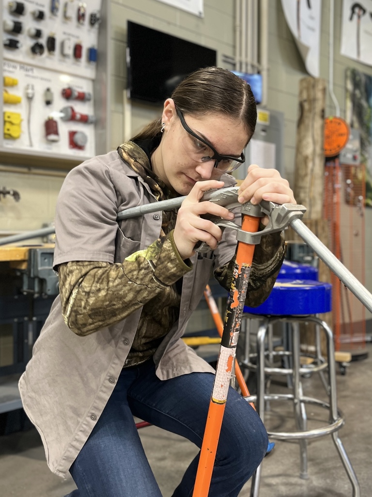 Electrical Occupations student wearing safety glasses uses a conduit bender to bend metal conduit in the classroom shop.