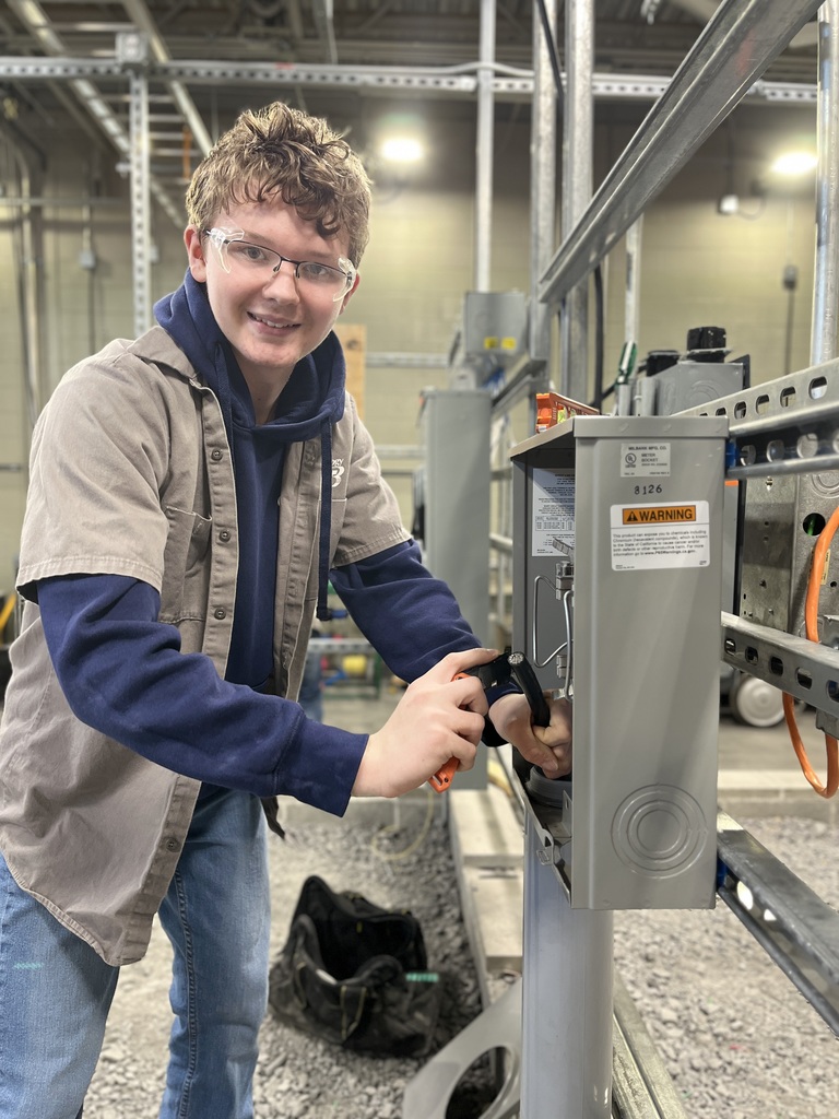 Electrical Occupations student wearing safety glasses smiles while using a hand tool on an open electrical panel mounted on a metal frame.