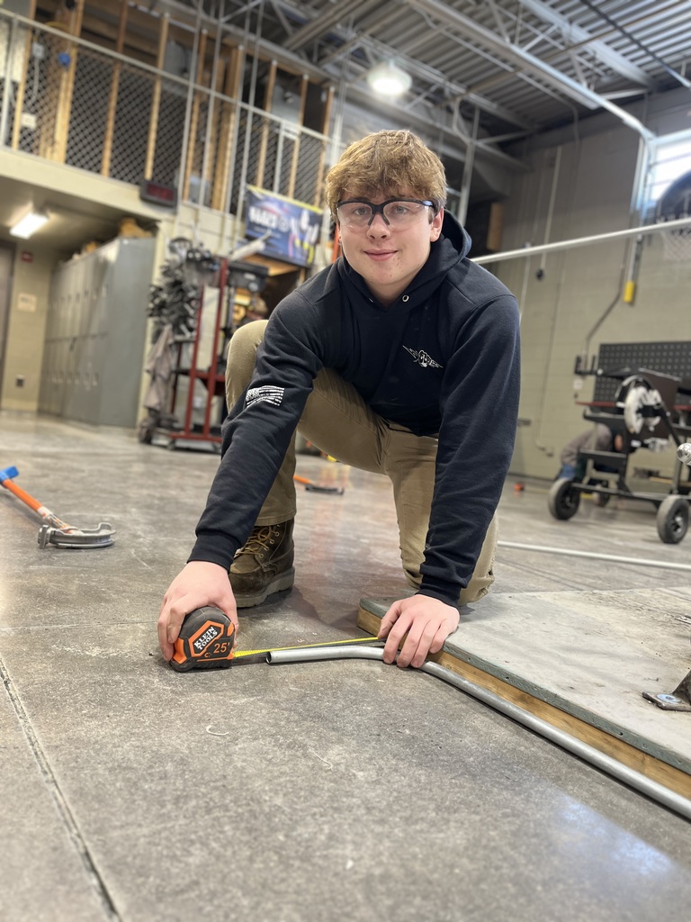 Electrical Occupations student kneels on the shop floor using a tape measure to measure a curved piece of electrical conduit.