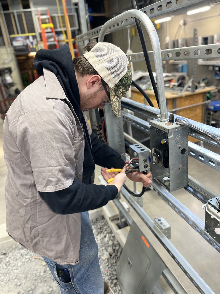 Electrical Occupations student wearing safety glasses uses hand tools to connect wiring inside a metal electrical box mounted on a metal frame.