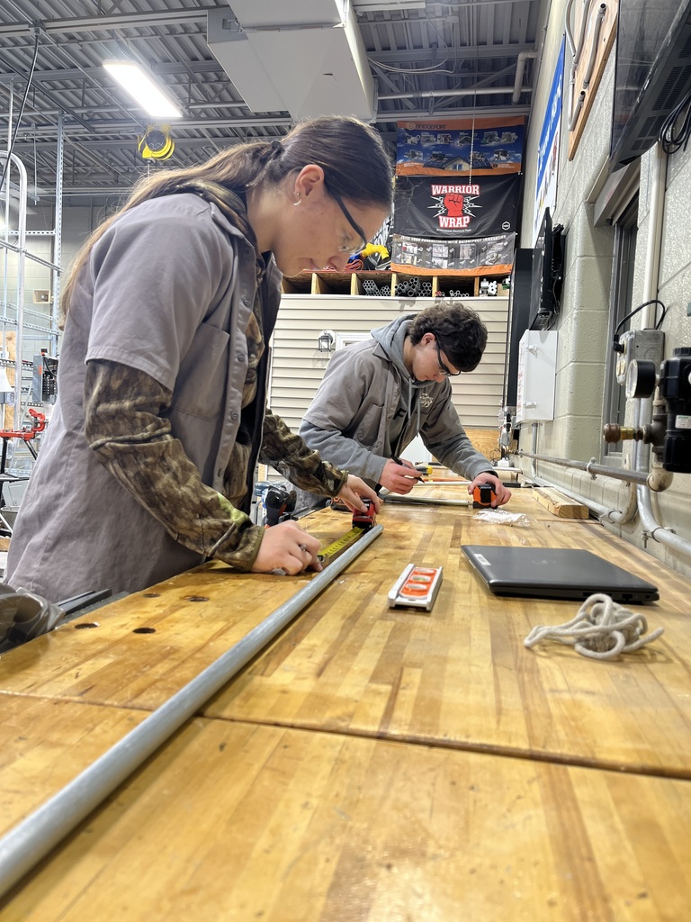 Two Electrical Occupations students measure and mark conduit on a wooden workbench in the shop.