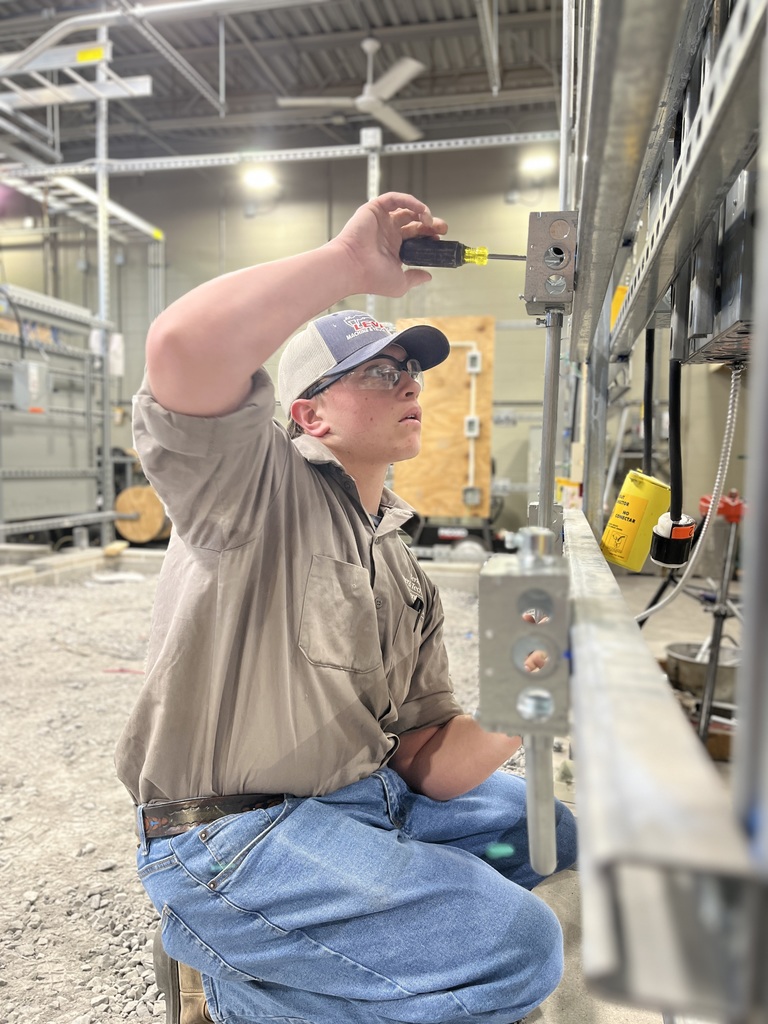 Electrical Occupations student wearing safety glasses kneels beside a metal frame and uses a screwdriver to work on a mounted electrical box.