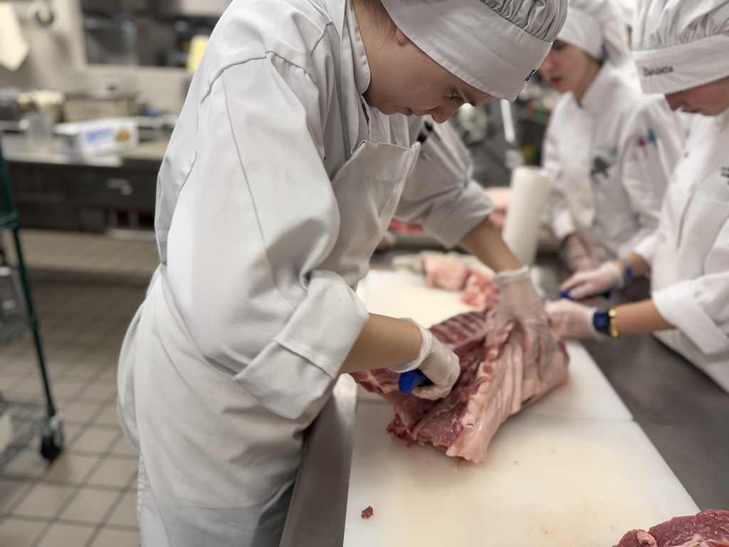 A Culinary student in a chef coat and hair covering uses a knife to cut along a pork rack on a cutting board while other students work beside the table in a commercial kitchen.