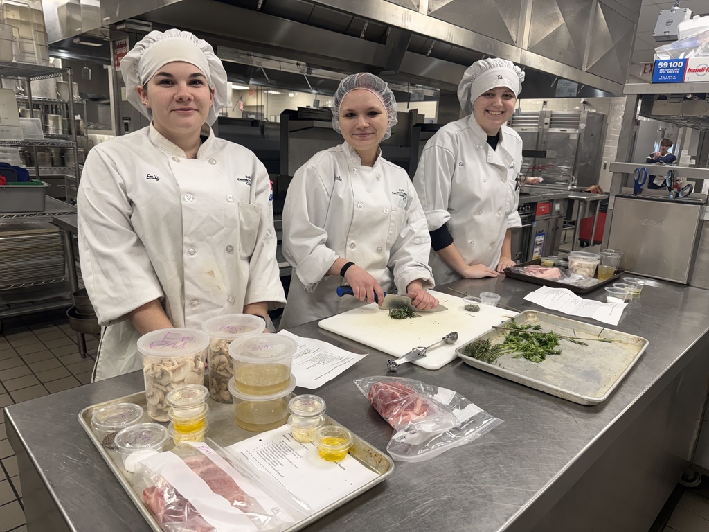 Three Culinary students in chef coats and hats stand behind a stainless-steel prep table in a commercial kitchen, with herbs, mushrooms, and pork portions arranged on trays and cutting boards.