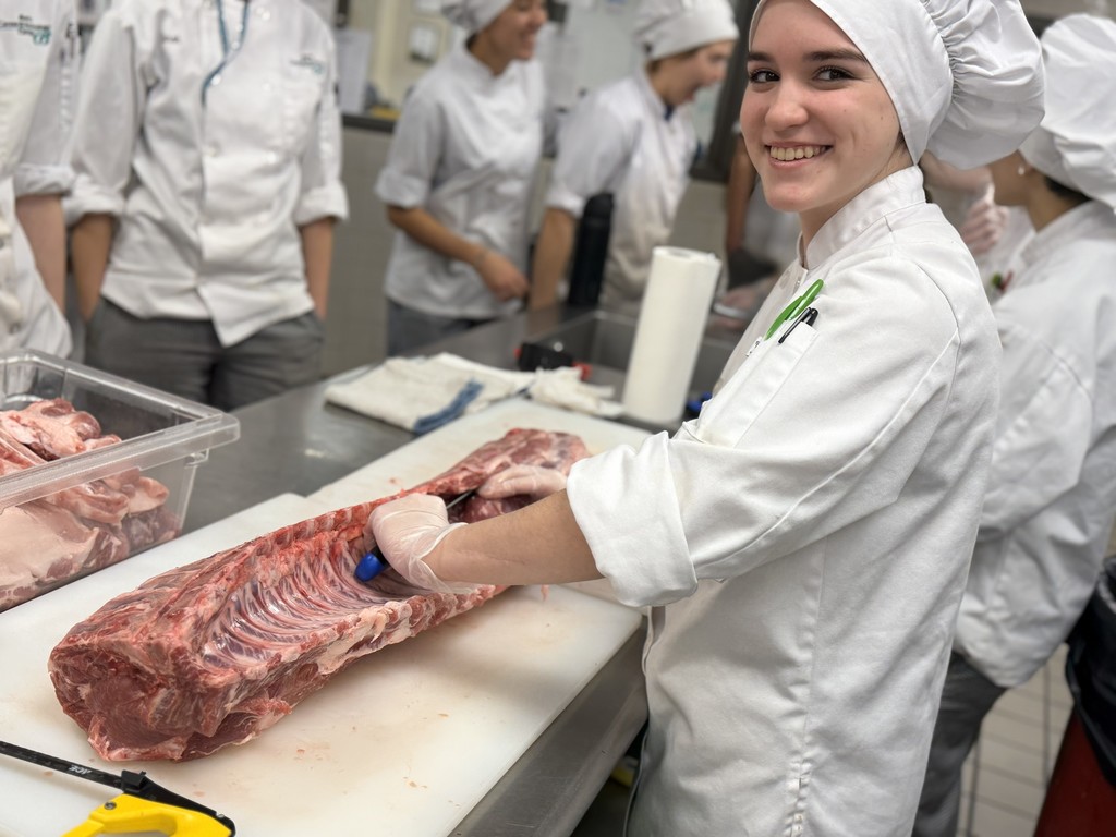 A Culinary student in a chef coat and hat smiles while trimming a large cut of pork on a cutting board at a stainless-steel worktable, with other students blurred in the background.