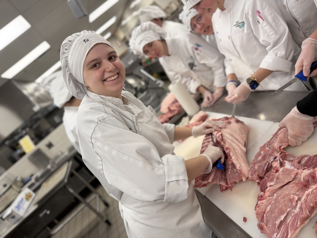 A Culinary student in a chef coat and hair covering smiles at the camera while cutting a pork rack on a cutting board, with classmates working nearby in the background.