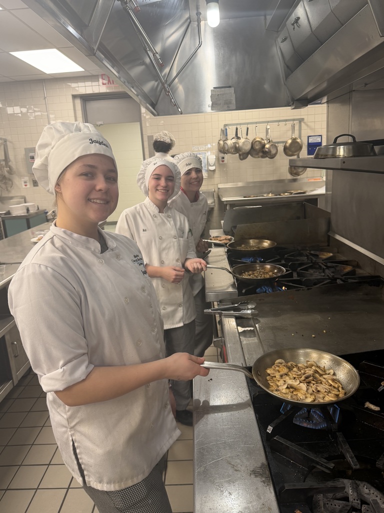 Three Culinary students in chef coats stand at a stove in a commercial kitchen, cooking mushrooms in a skillet over an open flame while smiling toward the camera.