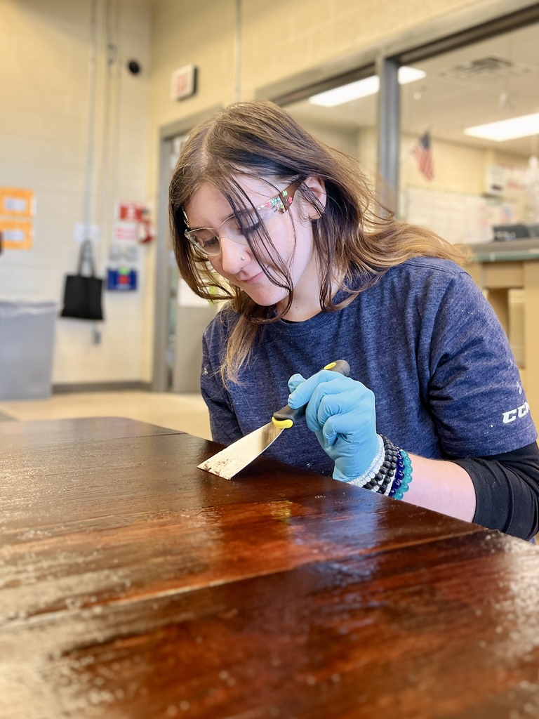 Student wearing gloves uses a putty knife to scrape and strip the old finish from a dark wooden tabletop.