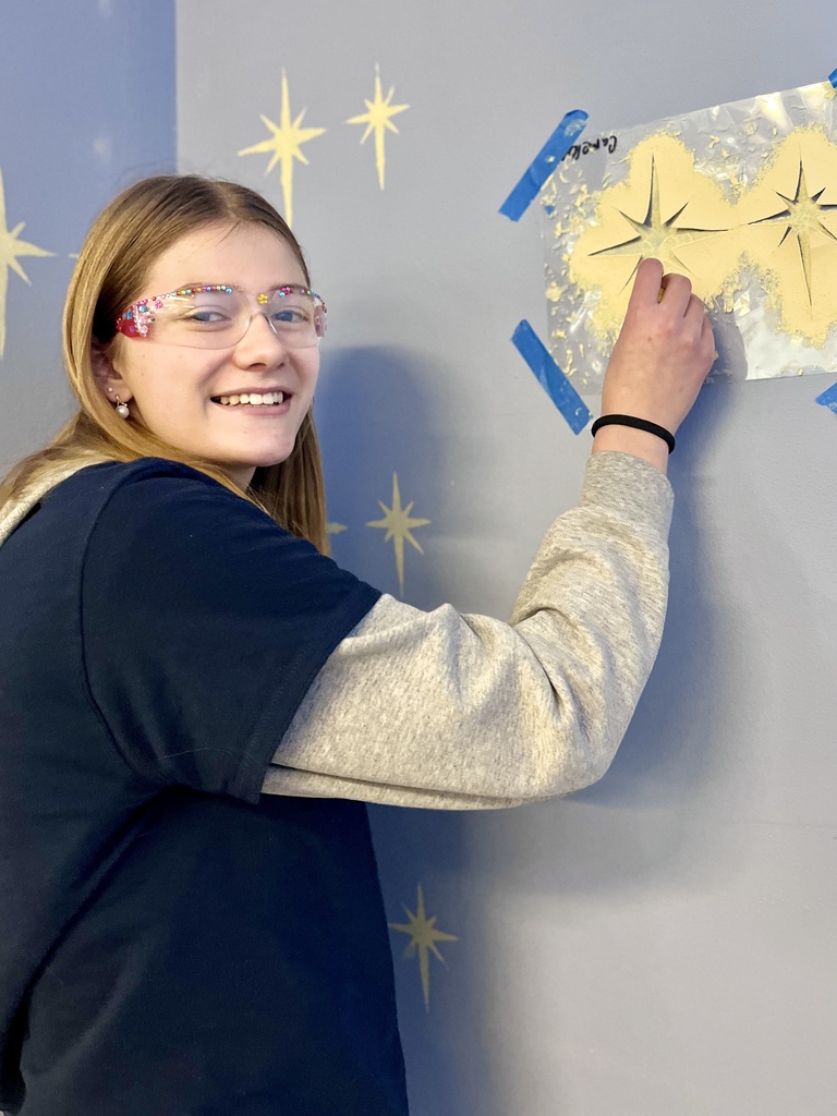 Smiling student wearing safety glasses uses a stencil on a blue wall, painting starburst shapes in a classroom project area.