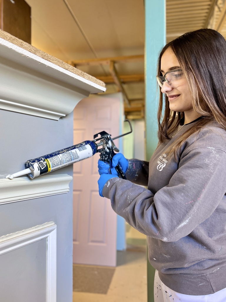 Student wearing safety glasses and blue gloves applies caulk with a caulking gun along trim detail on a painted wall section.
