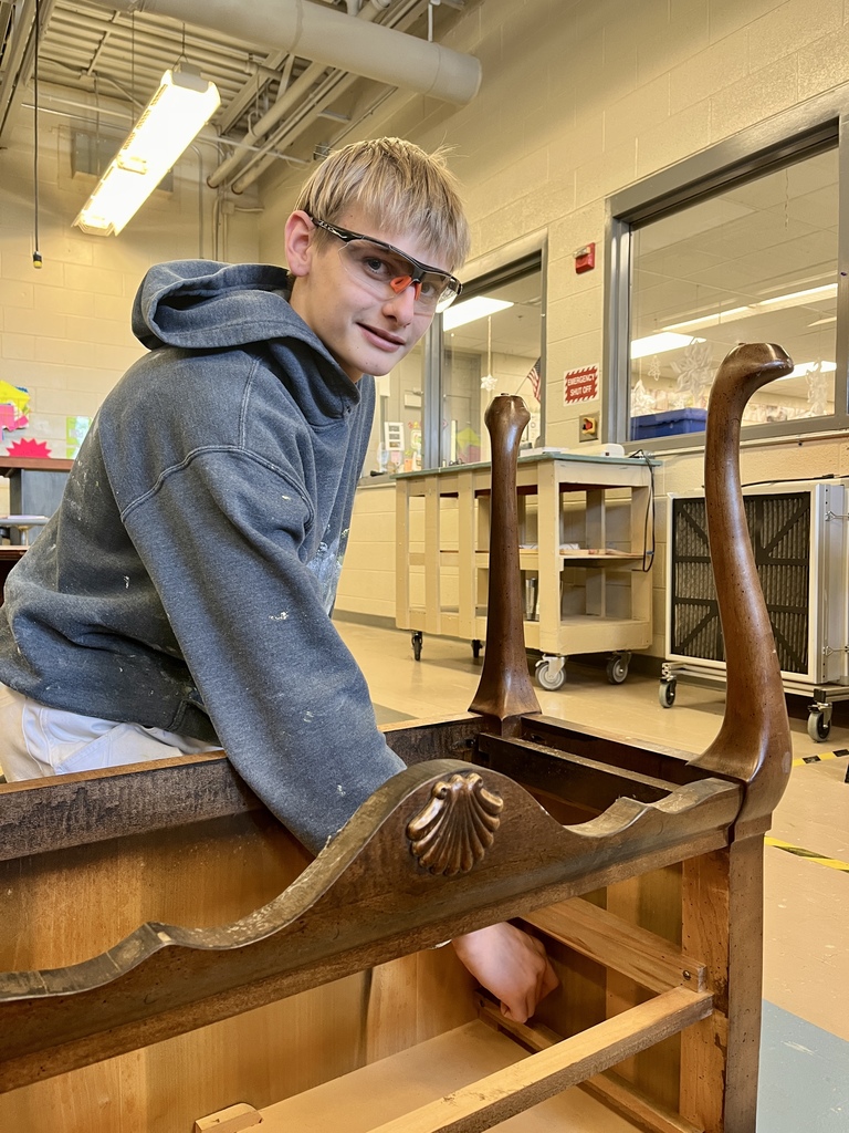 Student wearing safety glasses works on an upside-down wooden furniture piece, reaching inside the frame during the refinishing process.