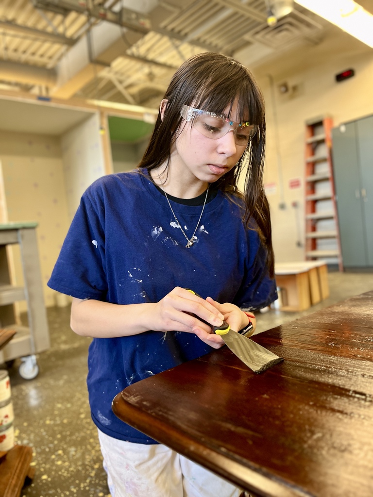 Student wearing safety glasses uses a putty knife to scrape and strip finish from the edge of a wooden tabletop in the workshop.