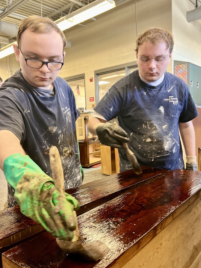 Two students wearing safety glasses and work gloves apply dark wood stain to a tabletop using paintbrushes.