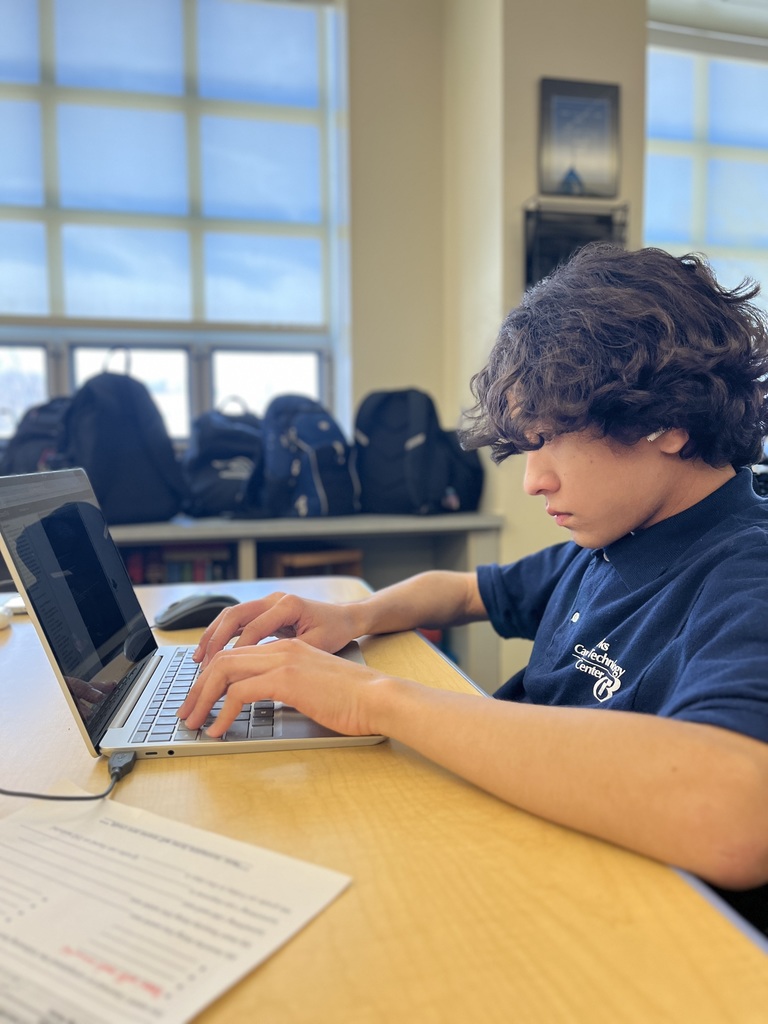 Student wearing a Berks Career & Technology Center polo works on a laptop at a classroom desk, focused on typing while a worksheet sits nearby. Backpacks line the windowsill behind him.
