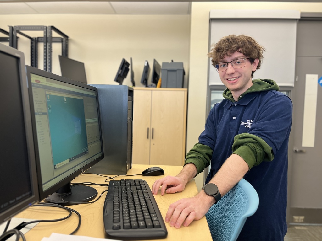Student in a BCTC polo sits at a desktop computer workstation in a lab, smiling while using a keyboard and mouse. Multiple computer monitors and lab equipment are visible in the background.