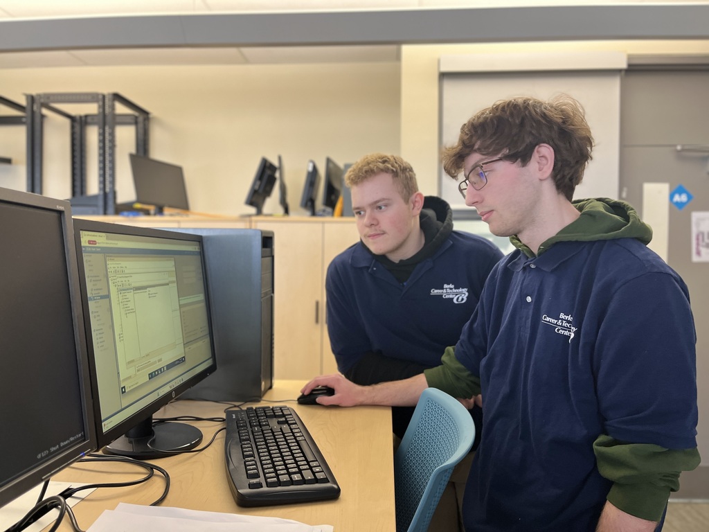 Two BCTC students stand and sit side by side at a computer workstation, looking at a monitor together while one student uses the mouse. Additional computer stations and lab storage cabinets are visible in the room.