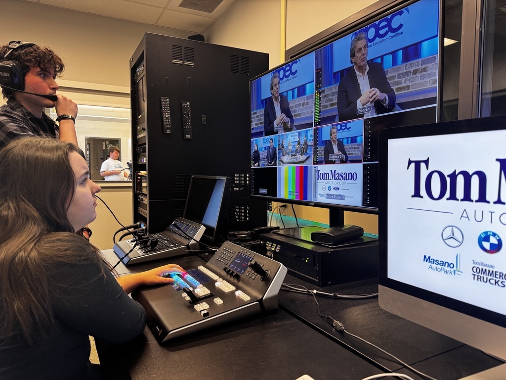 Two students in a video production control room wear headsets while monitoring a multi-screen display showing a studio interview in progress. One student uses a production switcher with illuminated buttons as camera angles and graphics appear on the screens.