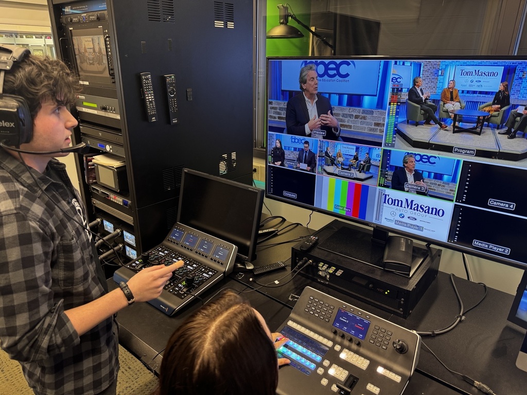 Student wearing a headset operates a video switcher in a control room while another student manages a lighting and camera control panel. Multiple monitors display a live studio interview set with three people seated on stage in front of BBEC and Tom Masano Auto Group signage.