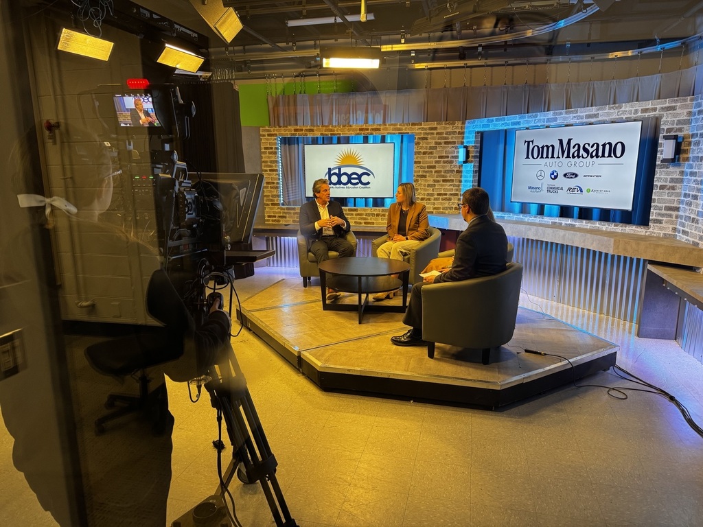 View through a studio window shows a three-person interview set with professional lighting, cameras, and a backdrop featuring BBEC and Tom Masano Auto Group branding. A camera operator films the seated speakers around a small table.