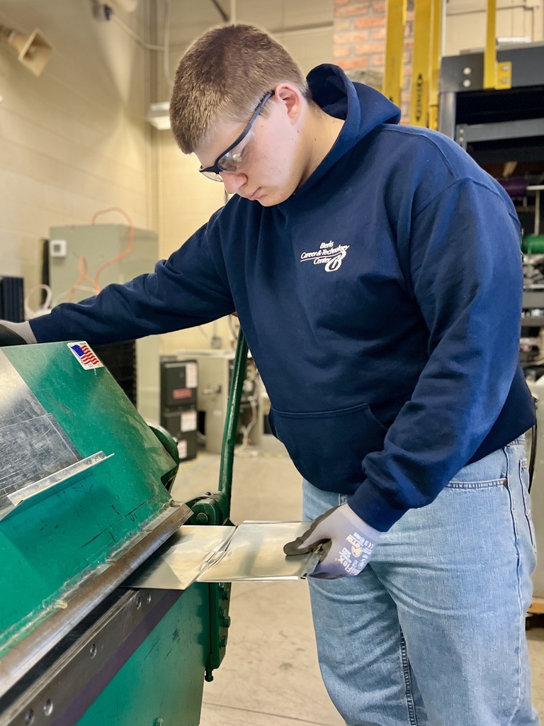 A student in a hoodie, safety glasses, and gloves operates a large green sheet metal brake to bend refurbished metal into a precise angle for HVAC duct components.