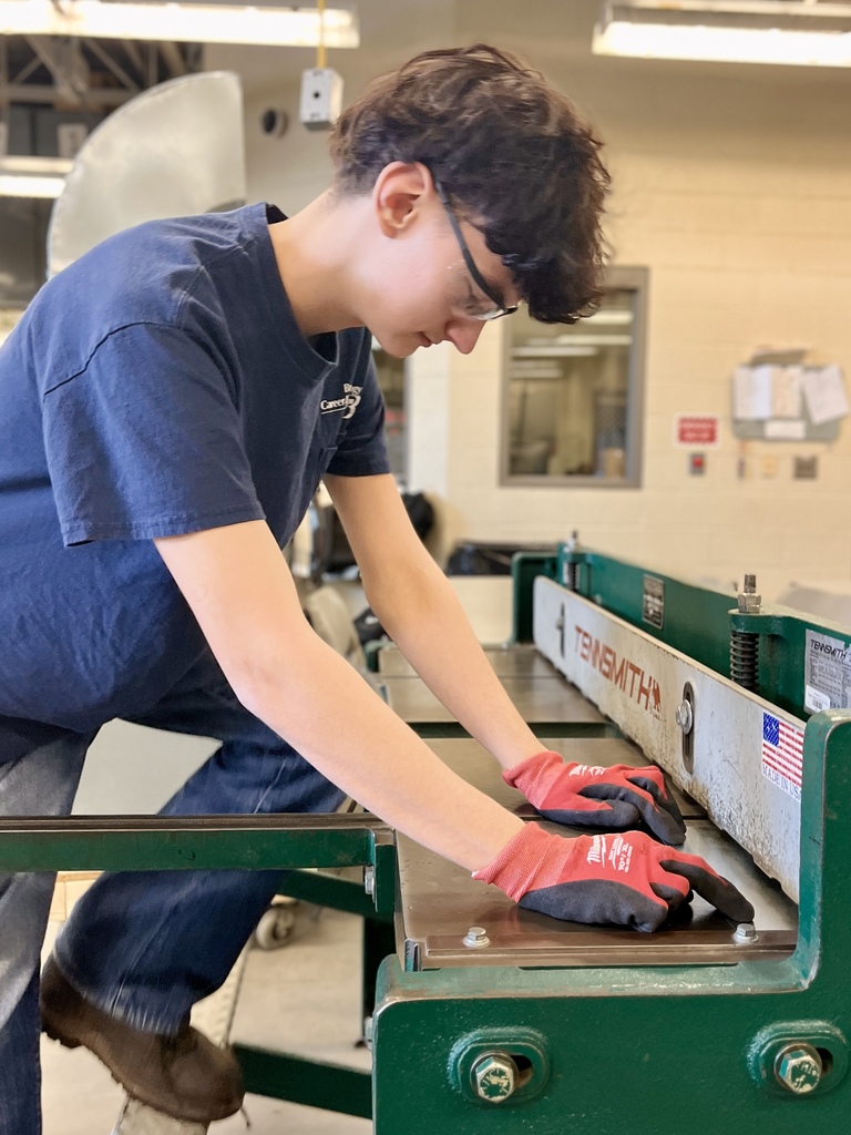 A student in safety glasses and red gloves guides a piece of refurbished sheet metal through a green metalworking machine to bend it into shape for ductwork.