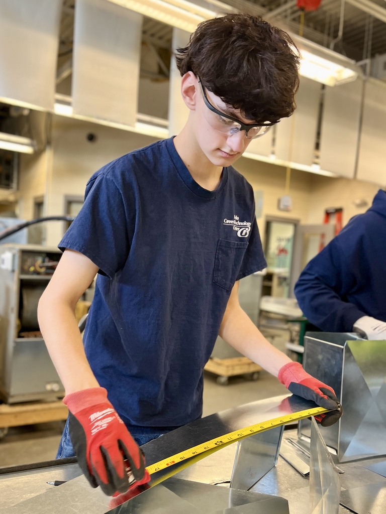 A student wearing safety glasses and red gloves measures a piece of refurbished sheet metal with a tape measure to ensure accurate sizing for duct elbow fabrication.