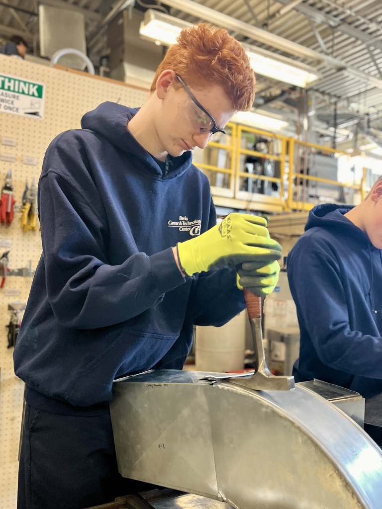 A student wearing safety glasses and bright green gloves carefully shapes a curved section of refurbished sheet metal using a forming tool to create a duct elbow in the HVAC shop.