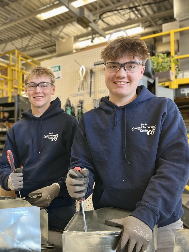 Two HVAC/Refrigeration students wearing safety glasses, gloves, and Berks Career & Technology Center hoodies use hand tools to fasten and shape refurbished sheet metal duct pieces at a workbench in a shop.