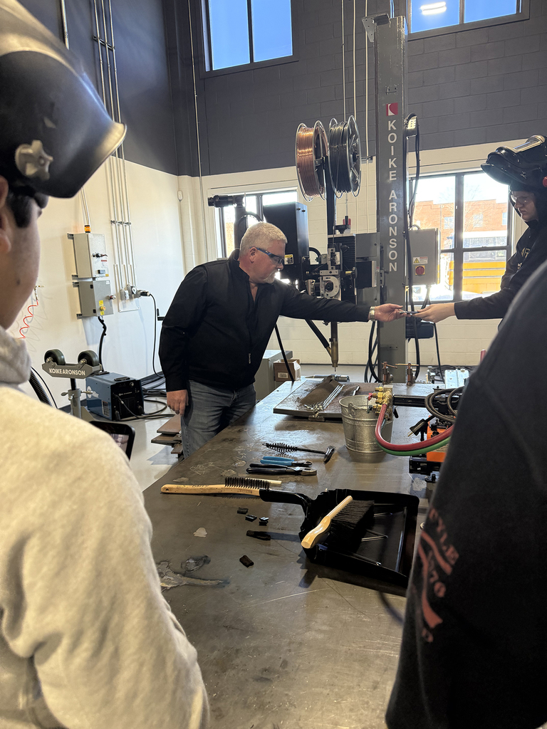 An instructor hands a welding sample or tool across a large metal worktable to a student during a hands-on demonstration.