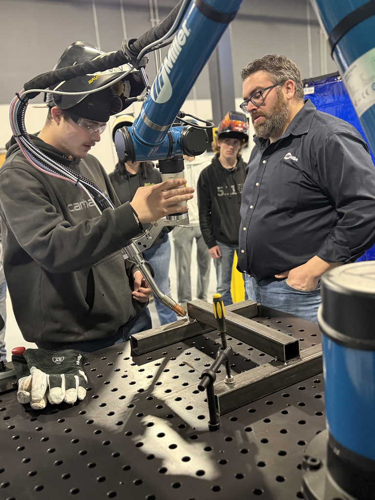 A student operates a robotic welding arm under the supervision of an industry representative, with other students watching closely in the background.